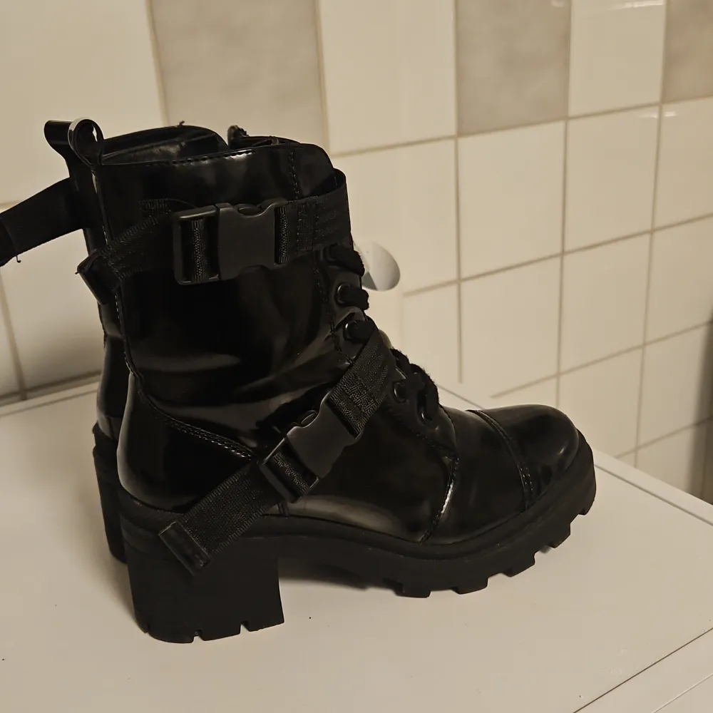 Säljer ett par svarta, glansiga boots i lack med både snörning spännen. De har en rejäl sula som ger bra grepp och en edgy look. Perfekta för höst och vinter när du vill ha något som både är snyggt och praktiskt. Passar till både jeans och k. Skor.