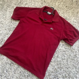 Vinröd pikétröja från Lacoste - Säljer en snygg vinröd pikétröja från Lacoste. Den har en klassisk krage och korta ärmar, perfekt för både vardag och lite finare tillfällen. Tröjan är i bomull och har den ikoniska krokodilloggan broderad på bröstet. Passar perfekt till både jeans och chinos!
