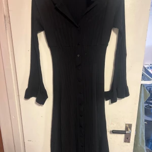 Svart långklänning med knappar - Säljer en elegant svart långklänning med knappar hela vägen ner. Klänningen har långa ärmar och en krage som ger en stilren look. Perfekt för höst och vinter, och kan kläs upp eller ner beroende på tillfälle.