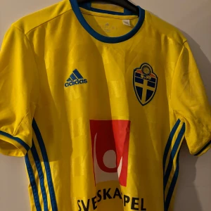 Sverigetröja - Säljer en gul fotbollströja från Adidas med blå detaljer och Sveriges landslagsemblem på bröstet. Tröjan har korta ärmar och blå ränder längs sidorna. Perfekt för fotbollsfans som vill visa sitt stöd för Sverige! 🇸🇪