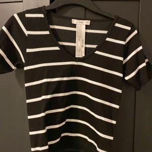 Svart och vit randig topp från MNG - Säljer en snygg svart och vit randig topp från MNG. Toppen har en ribbad struktur och en v-ringad hals. Den är kortärmad och passar perfekt för en stilren look. Perfekt för dig som gillar klassiska mönster! Aldrig använd