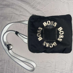Hugo boss axelväska - Snygg svart axelväska från Hugo Boss med ett stilrent tryck av märkesnamnet i cirkel på framsidan. Väskan har en justerbar axelrem i vitt och svart samt en praktisk dragkedja upptill. Perfekt för att bära med sig det nödvändigaste med stil. Pris går att diskutera