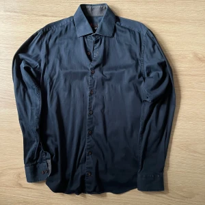 Svart skjorta från Dressmann - Snygg svart skjorta från Dressmann i storlek M. Skjortan har långa ärmar och knappar framtill. Perfekt för en stilren look.