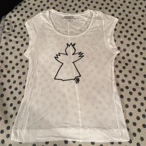 Vit t-shirt från By Malene Birger - Säljer en vit t-shirt från By Malene Birger med ett unikt svart tryck av en figur på framsidan. T-shirten är ärmlös och har en rund halsringning. Perfekt för en avslappnad stil🕊️