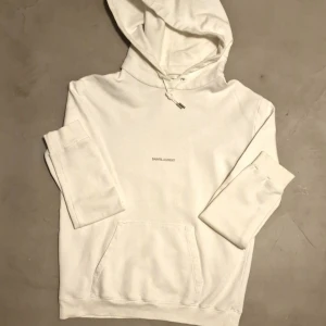Vit hoodie från Saint Laurent - Säljer en stilren vit hoodie från Saint Laurent| skick:8/10| str:M Längd 70 cm , bredd under arm ca 57cm | nypris: 8 000kr+ vårat pris 1 699kr| tveka inte på att höra av dig för fler bilder eller frågor!🤩😁