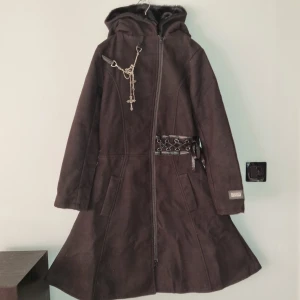 Svart kappa från Black Alchemy - Snygg svart kappa från Black Alchemy med huva och dragkedja framtill. Den har en cool snörning på ryggen och kedjedetaljer i midjan. Perfekt för en edgy look!