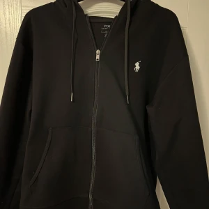 Svart hoodie från Polo Ralph Lauren - Säljer en stilren svart hoodie från Polo Ralph Lauren med dragkedja och klassisk logga på bröstet. Tröjan har en bekväm passform och praktiska fickor framtill. Perfekt för en avslappnad look.