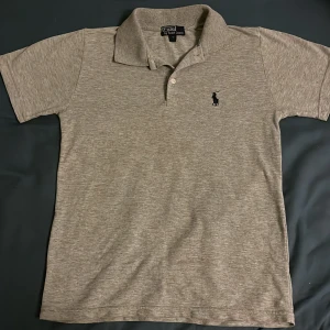 Grå pikétröja från Ralph Lauren - Säljer en klassisk grå pikétröja från Ralph Lauren med den ikoniska broderade loggan på bröstet. Tröjan har korta ärmar och en stilren krage med knappar. Pris kan diskuteras.