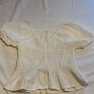 Vit blus från Kappahl - Charmig vit blus från Kappahl med korta puffärmar och volangdetalj vid nederkanten. Blusen har en smockad design som ger en fin struktur och passform. Perfekt för en somrig look!