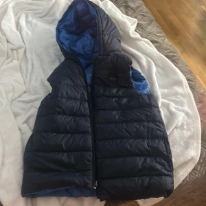 Blå och svart dunväst från BOSS - Säljer en snygg och vändbar dunväst från BOSS i blått och svart. Den är även två sidig. Västen har en praktisk dragkedja och en huva för extra skydd. Perfekt för kyligare dagar och ger en stilren look.