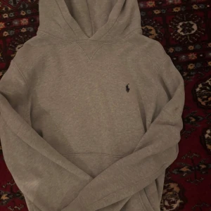 Grå hoodie från Ralph Lauren - Säljer en stilren grå hoodie från Ralph Lauren med den klassiska broderade loggan på bröstet. Perfekt för en avslappnad look. Hoodien har en bekväm passform och är tillverkad i mjukt material.
