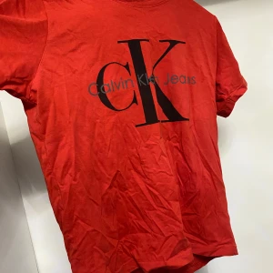 Röd t-shirt från Calvin Klein - Snygg röd t-shirt från Calvin Klein med stort CK-logotyptryck på framsidan. Perfekt för en avslappnad stil. T-shirten har en klassisk rund hals och korta ärmar.