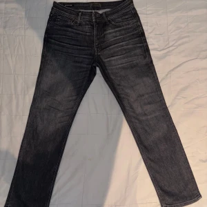 Svarta jeansbyxor - Snygga svarta Jack & Jones jeansbyxor med en klassisk straight passform. Modellen på Jeansen är JJCLARK. De har en dragkedja och knappar framtill. Perfekta för en stilren look.