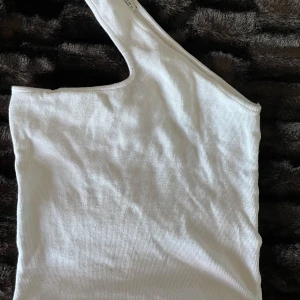 Vit oneshoulder topp - Snygg vit oneshoulder topp i ribbat material. Perfekt för en stilren look med en unik asymmetrisk design. Passar bra till både jeans och kjol.
