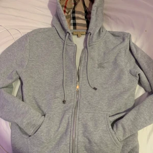 Grå huvtröja från Burberry - Säljer en stilren grå huvtröja från Burberry med dragkedja och klassiskt rutigt mönster i fodret. Tröjan har en justerbar huva och är tillverkad i mjukt material för extra komfort. Perfekt för en avslappnad look.skriv för mer info. den är xl men är som en medium