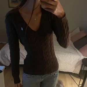 Brun stickad tröja med v-ringning från Ralph lauren  - Säljer en snygg brun stickad tröja med v-ringning och kabelstickat mönster. Tröjan har långa ärmar och en liten logga på bröstet. Perfekt för kyligare dagar!