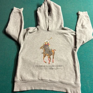 Grå hoodie från Ralph Lauren - Säljer en grå hoodie från Ralph Lauren med ett coolt tryck av en polospelande björn på framsidan. Tröjan har långa ärmar och en klassisk huva. Perfekt för en avslappnad stil. 🐻 lite av polo bear loggan har åkt bort i tvätten, säljer denna för att den är för liten