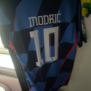 Blå och svart fotbollströja med Modric 10 - Snygg fotbollströja i blått och svart med Modric och nummer 10 på ryggen. Tröjan har ett schackrutigt mönster och korta ärmar. Perfekt för fotbollsfans! Storlek M