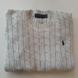 Grå stickad tröja från Polo Ralph Lauren - Säljer denna grymt fina kabelstickade tröjan från Ralph Lauren, ljusgrå /greige med marinblå logga. Stl M men lite liten i storlek så passar även en S beroende på hur du vill att den skall sitta, se mått. Självklart äkta! Riktigt fint skick, mjuk och härlig kvalitet med stretch. Mått: Armhåla till armhåla 53cm, bredd längst ner 46cm, längd från mitt på axeln 62cm. Ordinarie pris 2500, så denna får du till ett riktigt fint pris! 