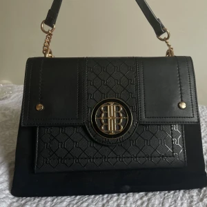 Svart axelväska från River Island - Snygg svart axelväska från River Island med gulddetaljer och ett elegant mönster på framsidan. Väskan har en kedja som axelrem och ett stilrent emblem i mitten. Perfekt för att ge din outfit en lyxig touch.
