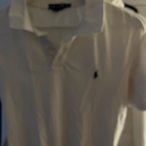 Vit pikétröja från Ralph Lauren - Säljer en klassisk vit pikétröja från Ralph Lauren med den ikoniska loggan broderad på bröstet. Tröjan har en krage och knappar framtill, perfekt för en stilren look. Passar både till vardags och mer uppklädda tillfällen.