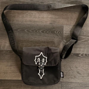 Trapstar väska rep - Snygg svart axelväska med ett unikt spänne framtill och justerbar axelrem. Väskan har en stilren design med vita detaljer som ger den en cool look. Perfekt för att bära dina nödvändigheter med stil. 