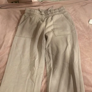 Beige mjukisbyxor med elastisk midja - Säljer ett par bekväma beige mjukisbyxor med elastisk midja och snörning. Byxorna har stora fickor fram och en bakficka, perfekta för en avslappnad stil. Materialet är mjukt och skönt, idealiskt för mysiga dagar hemma.