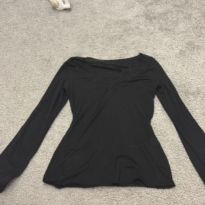 Svart långärmad topp med spetsdetaljer - Snygg svart långärmad topp med v-ringad spetsdetalj vid halsen och spets vid ärmsluten. Perfekt för en stilren look. Passar bra till både jeans och kjol.