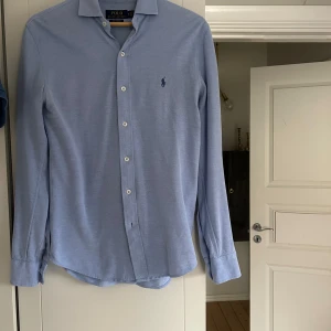 Blå skjorta från Ralph Lauren - Snygg blå skjorta från Ralph Lauren med klassisk krage och knappar framtill. Den har en broderad logotyp på bröstet och är tillverkad i ett bekvämt material. Perfekt för en stilren look. På lappen står det att skjortan är xs men jag är runt s-m men den passar mig väldigt bra
