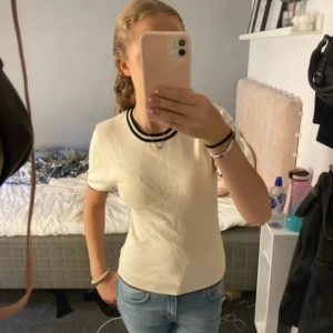 Vit kortärmad topp med svarta detaljer - Säljer en stilren vit topp med svarta ränder vid halsringningen. Toppen är kortärmad och har en tight passform, perfekt för en enkel och snygg look.