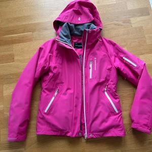 Rosa skidjacka från Peak Performance  - Säljer en snygg rosa skidjacka från Peak Performance. Jackan har en justerbar huva och flera praktiska fickor med dragkedjor samt ventilation i armhålan. Perfekt för blåsiga och kalla dagar då den är lätt paddad. Den är sparsamt använd, har en bekväm passform med stretch material, värmelås i midjan och tejpade sömmar. Djur och rökfri. Strl. M