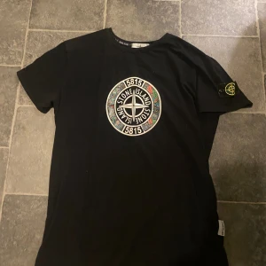 Svart t-shirt från Stone Island - Säljer en svart t-shirt från Stone Island med det ikoniska märket broderat på ärmen och ett stort tryck på framsidan. Perfekt för en stilren look.