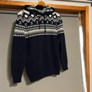 Mönstrad stickad hoodie i blått och vitt - Säljer en snygg stickad hoodie i mörkblått med ett stilrent mönster i vitt och grått. Tröjan har långa ärmar och en praktisk huva med snörning. Perfekt för kyliga dagar!