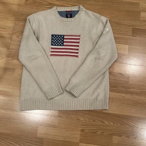 Beige tröja från GANT med amerikansk flagga - Säljer en beige tröja från GANT med en stor amerikansk flagga på framsidan. Tröjan är tillverkad i 100% bomull och har långa ärmar med ribbade muddar. Perfekt för en avslappnad stil.