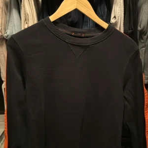 Svart långärmad tröja med orange detaljer från Parajumpers - Snygg svart långärmad tröja från Parajumpers med coola orange detaljer längs ärmarna. Tröjan har en rund halsringning och ett märke på ärmen. Perfekt för en stilren look.