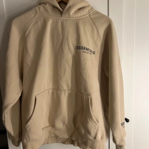 Beige hoodie från Essentials Fear of God - Säljer en stilren beige hoodie från Essentials Fear of God. Den har en klassisk design med känguruficka och tryck på bröstet och ärmen. Perfekt för en avslappnad look.