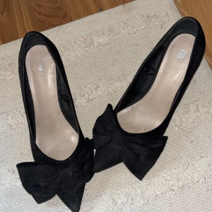 Svarta pumps med rosett - Svarta klackar i mocka med en stor dekorativ rosett framtill. Skorna har en spetsig tå och en hög klack, väldigt stilrena och extremt snygga, säljer då jag redan har andra. Inte använda på länge därav lite dammiga men går att fräscha upp enkelt!
