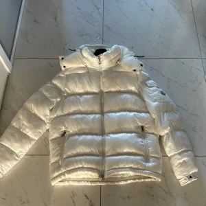  Moncler jacka vit - Säljer en snygg vit dunjacka från Moncler med hög krage och huva. Jackan har en dragkedja framtill och praktiska fickor med dragkedja. Den är långärmad och har en broderad logotyp på ärmen. Perfekt för kalla dagar!
