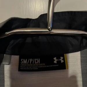 Grå och vit jacka från Under Armour - Säljer en grå och vit jacka från Under Armour med halv dragkedja och hög krage. Jackan har en loose passform och är perfekt för träning. Den har långa ärmar och en stilren design med logotyp på bröstet.