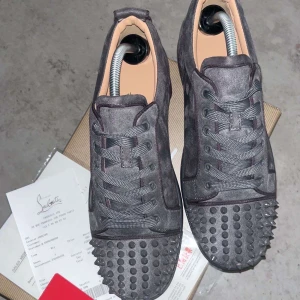 Grå sneakers med nitar från Louboutin - Snygga grå sneakers från Louboutin med nitar på tån och en ikonisk röd sula. Skorna har snörning och är tillverkade i mocka, vilket ger en lyxig känsla. Perfekta för att ge din outfit en edgy touch.