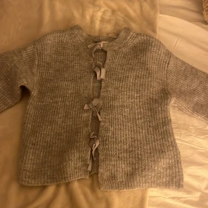 Grå stickad kofta med knytband - Mysig grå stickad kofta med knytband. Koftan har långa ärmar och en rund halsringning. Köpt från Gina använd en gång storlek 146-152