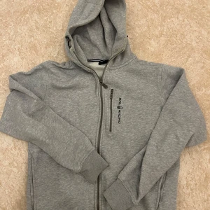 Sail Racing zip hoodie - Säljer en grå zip hoodie från Sail Racing i mycket bra skick. Den har en dragkedja framtill och en praktisk huva. Perfekt för kyliga dagar eller som ett extra lager. Tröjan är mjuk och bekväm, idealisk för både vardag och fritid. Säljer den eftersom den har ingen användning längre och har inte gjort det på länge!