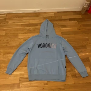Hoodrich hoodie ljusblå - Säljer en snygg ljusblå hoodie från Hoodrich i storlek M. Den har en stor logga på bröstet och en praktisk ficka framtill. Perfekt för en avslappnad stil och passar bra till både vardag och chill. Huvan har dragsko för justerbar passform.