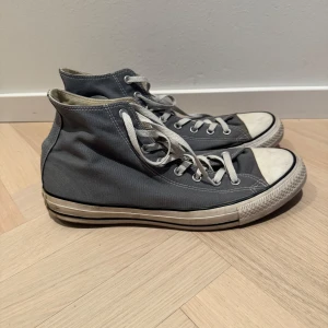 Grå Converse All Star sneakers - Säljer ett par klassiska grå Converse All Star sneakers i bra skick. De har den ikoniska höga modellen med vita snören och gummisula. Perfekta för vardagsbruk och ger en avslappnad stil. Lite slitna men fortfarande snygga och bekväma.