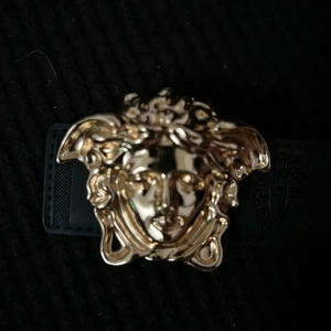 Svart skärp med guldspänne från Versace - Säljer ett snyggt svart skärp från Versace med ett stort, detaljerat guldspänne i form av Medusa. Skärpet är i skinn och har en lyxig känsla, perfekt för att lyfta vilken outfit som helst. Passar både till fest och vardag. 🖤✨