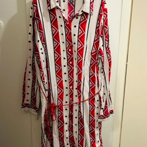 Mönstrad klänning från Rainbow - Säljer en färgglad klänning från Rainbow i storlek XL. Den har ett coolt geometriskt mönster i rött, svart och vitt med knappar framtill och ett knytband i midjan. Perfekt för en avslappnad dag eller en sommarfest! 🌞