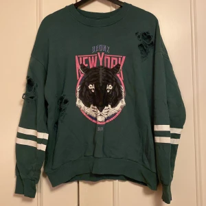 Grön sweatshirt från Gina Tricot - Cool grön sweatshirt från Gina Tricot med ett stort tryck av en svart panter och texten 'Bronx New York'. Tröjan har slitna detaljer på ärmarna och vita ränder. Perfekt för en avslappnad stil!