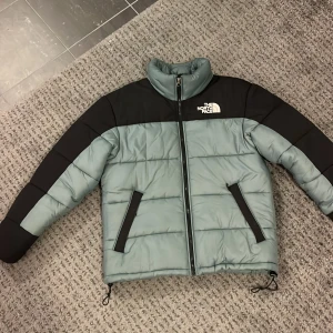 Grå och svart pufferjacka från The North Face - Säljer en snygg och varm pufferjacka från The North Face i grå/grön och svart i storlek M. Jackan har en dragkedja framtill och är perfekt för kalla dagar. Den är i mycket bra skick och har långa ärmar. Perfekt för höst och vinter! 