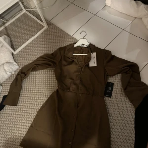 Brun skjortklänning i satin från NA-KD - Oanvänd brun skjortklänning i satin från NA-KD i storlek EU 34. Klänningen är i satin och har långa ärmar med knappar framtill. 