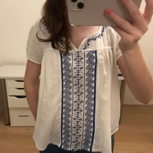 Vit blus med blå broderier - Säljer en superfin vit blus med blå broderier framtill. Den har korta ärmar och en lös passform, perfekt för vår och sommar. Blusen är i mycket bra skick och passar både till vardags och festliga tillfällen.